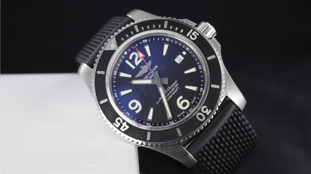 Breitling horloge met blauwe wijzerplaat en zwarte rubberen band half schuin liggend op wit blokje met zwarte achtergrond.