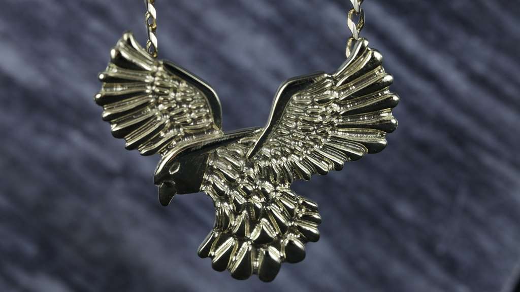 Geelgouden adelaar in 3D als hanger aan collier, vastgehouden aan 2 oogjes aan vleugels op blauwe ondergrond.