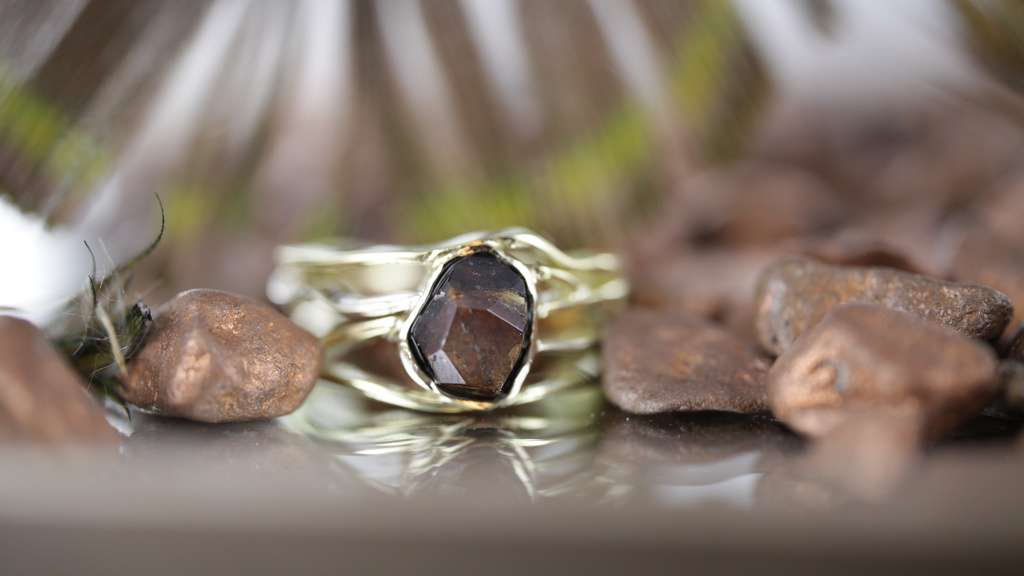 Geelgouden ring 4 banden met opengewerkte structuur, eigen bruine steen van klant in verwerkt op glazen ondergrond, bruine stenen rondom ring en met lichtstralen op achtergrond.