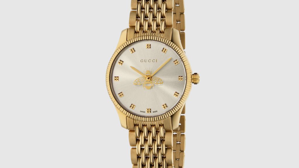 Gucci gold plated horloge, met goudkleurige horlogekast en band, en zilveren zongebrande wijzerplaat met gouden bij, vooraanzicht op licht grijze ondergrond.