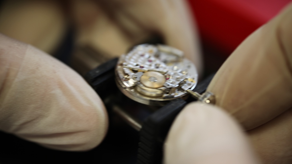 Horlogemaker draait aan onderdeel van Danish Design uurwerk met handschoenen aan en rode achtergrond.