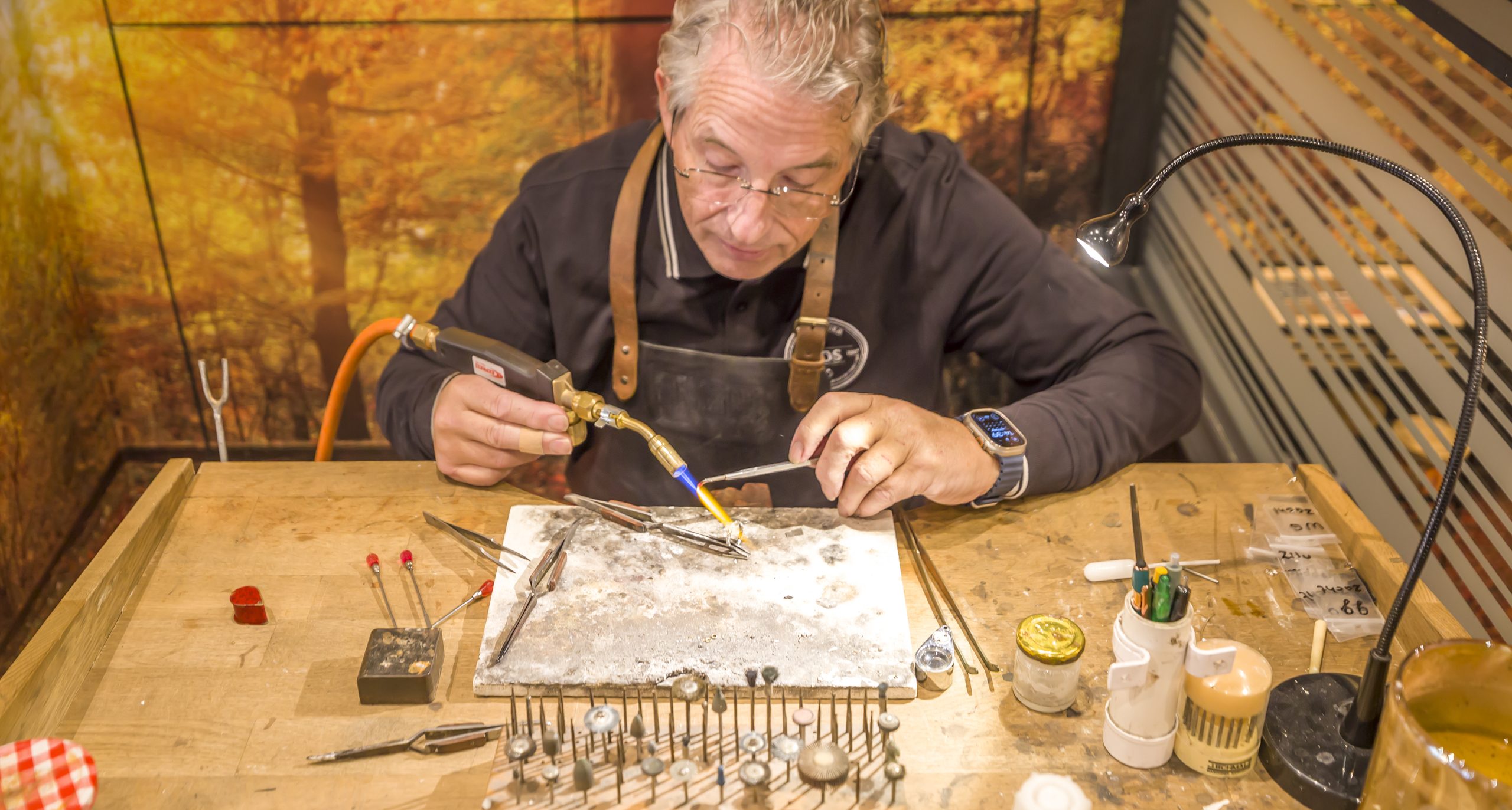 Juwelier Jos van Beek zit achter werkband en soldeert een ring met een brander achter werkbank.