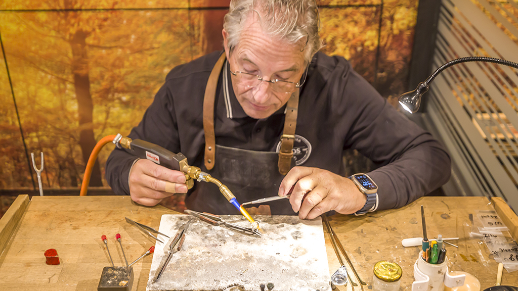 Goudsmid Jos van Beek achter werkbank, bezig met het solderen van een ring met een vlam.