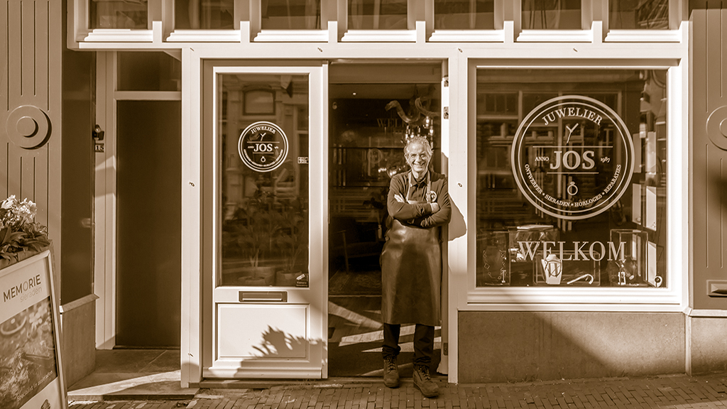 Goudsmid Jos van Beek staat voor winkel in opening met handen over elkaar, foto met sepia filter.