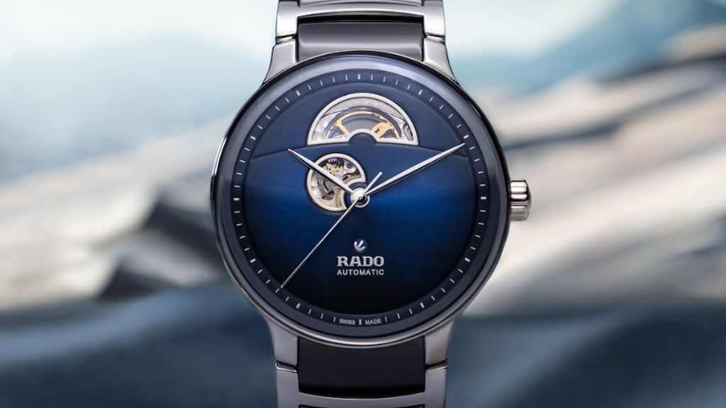 Stalen Rado horloge met blauwe wijzerplaat, deel van uurwerk zichtbaar en blauw keramieken en stalen band op bergachtige blauwe vage achtergrond.