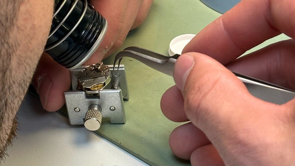 Uurwerkmaker repareert uurwerk met pincet en bekijkt met loep het geheel.