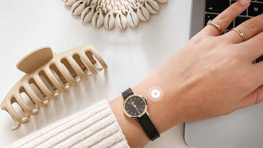 Danish Design horloge met gouden accenten en zwart leren band om pols van vrouw achter laptop met daarnaast haarclip en schelpen accesoire op witte ondergrond.